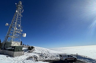 A telecommunications tower and a Strabag service vehicle on a snowy mountain summit above a sea of clouds.