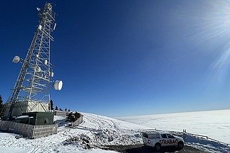 Ein Fernmeldeturm und ein Strabag-Dienstfahrzeug auf einem verschneiten Berggipfel über einem Wolkenmeer.