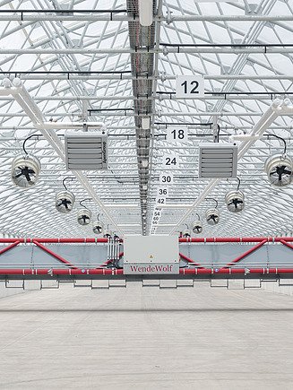 Front view of an automated sludge turning and conveying bridge inside a solar sludge drying hall. The equipment spans the full width of the hall and is used to loosen, turn, and transport sewage sludge during drying. Ventilation fans and heating units installed throughout the greenhouse structure support the solar drying process and optimize moisture removal.
