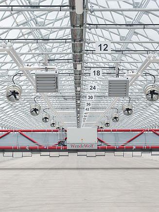 Frontalansicht eines automatischen Schlammwende- und Fördergeräts in einer Solartrocknungshalle. Das über die gesamte Hallenbreite reichende System sorgt für das Lockern, Wenden und Verteilen des Klärschlamms, während Ventilatoren und Heizgeräte den Trocknungsprozess unterstützen.