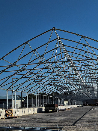 Bauphase einer solaren Klärschlammtrocknungsanlage, die einen großen offenen Betonsockel mit einer teilweise montierten Stahlrahmenkonstruktion unter blauem Himmel zeigt.