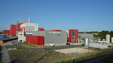 Diese Luftaufnahme zeigt die komplette Biomethanisierungsanlage mit ihren verschiedenen Gebäuden, Tanks und der Umgebung unter einem klaren blauen Himmel. An der rot-grauen Fassade ist der Schriftzug 