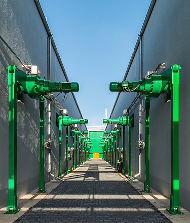 Das Bild zeigt einen symmetrischen Korridor zwischen zwei Industriegebäuden, der mit grünen, auf stabilen Stützen montierten mechanischen Einheiten gesäumt ist.